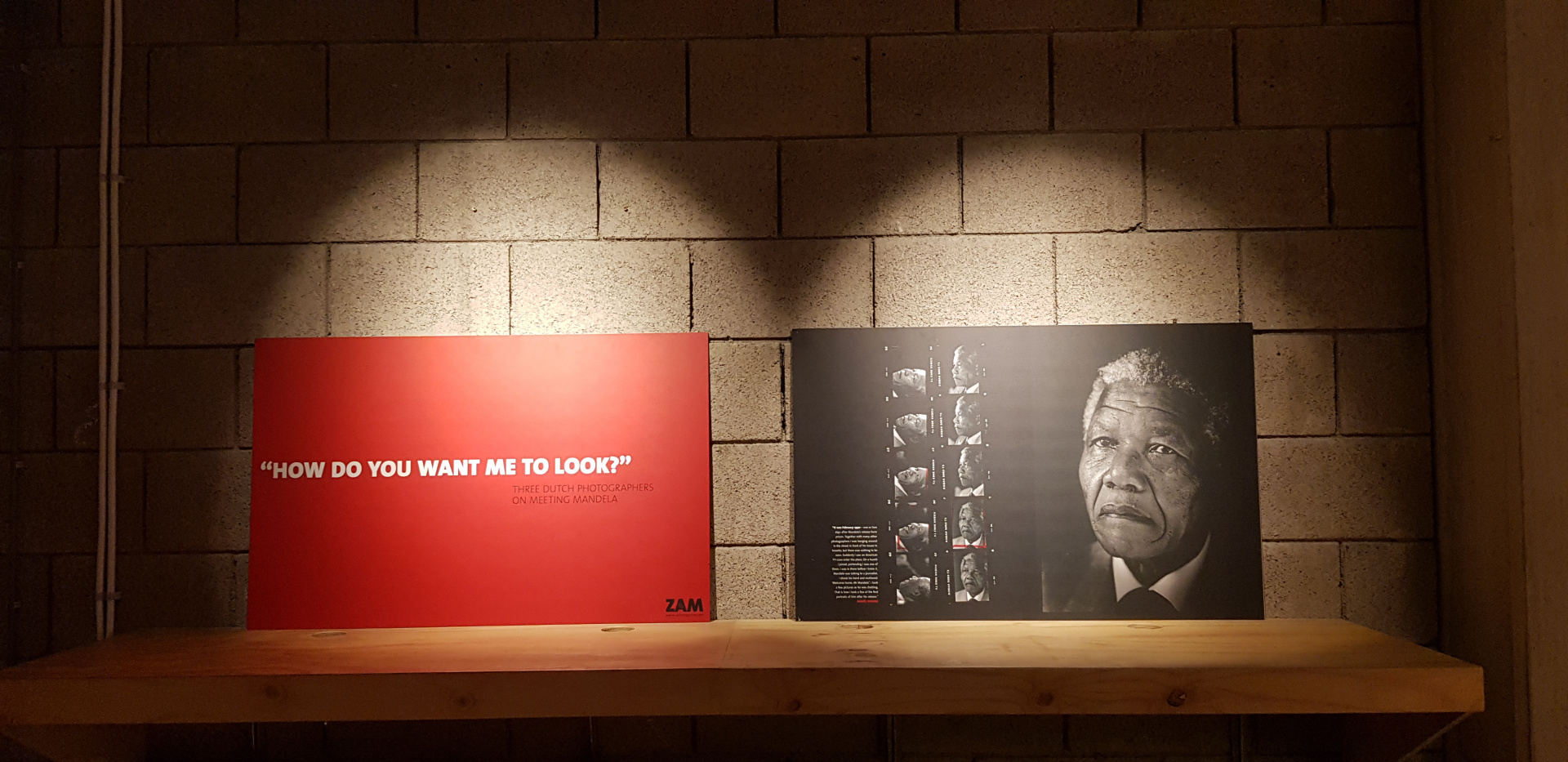 Hier zie je een m uur met 2 grote foto's die door een spotlight worden verlicht. Op de linkerfoto zie je een rood vlak met gecentreerd in het midden de tekst in witte kapitalen: 'How do you want me to look?'. Op de rechterfoto zie je aan de linkerkant 2 kolommen met kleine foto's en aan de rechterkant een grote foto van het gezicht van Nelson Mandela. 